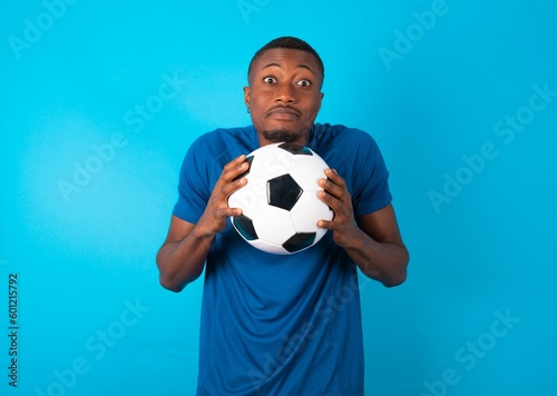 Happy Young man wearing sport T-shirt holding a ball over blue background keeps fists on cheeks smiles broadly and has positive expression being in good mood