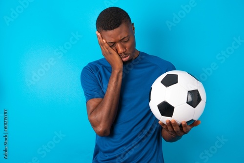 Sad lonely Young man wearing sport T-shirt holding a ball over blue background touches cheek with hand bites lower lip and gazes with displeasure. Bad emotions