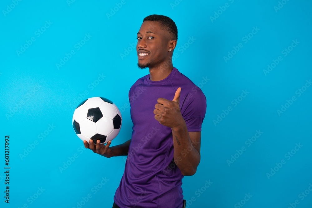 Man wearing purple T-shirt holding a ball over blue background giving thumb up gesture, good Job! Positive human emotion facial expression body language. Man wearing purple T-shirt holding a ball over blue background giving thumb up gesture, good Job! Positive human emotion facial expression body language.