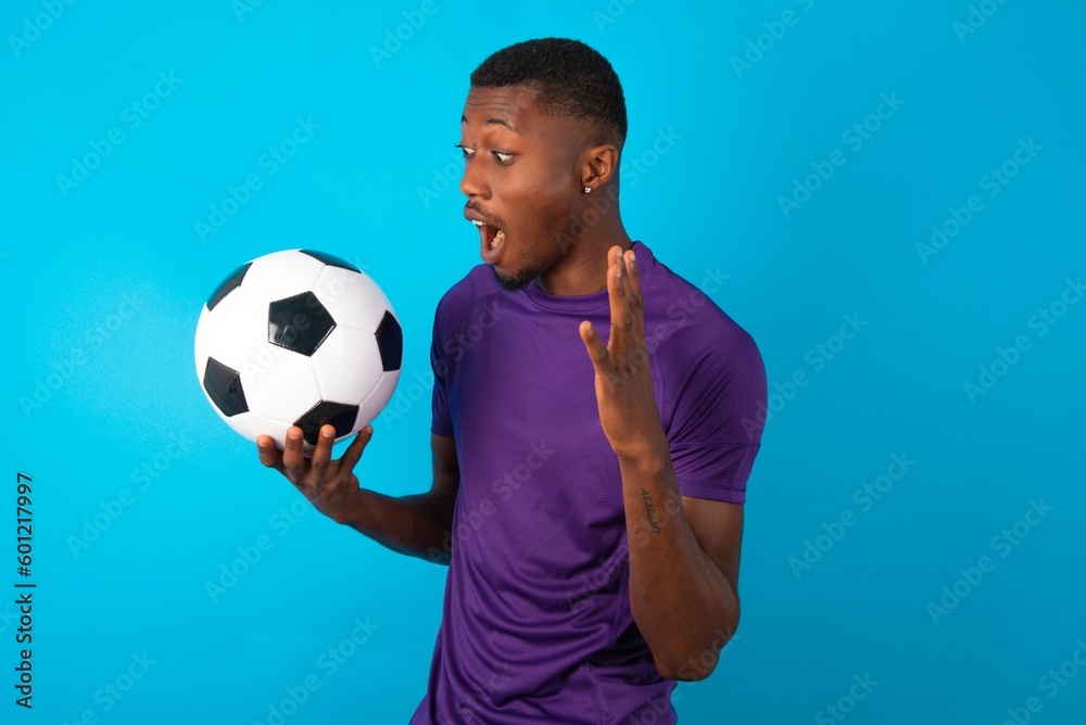Man wearing purple T-shirt holding a ball over blue background with shocked facial expression, holding hands near face, screaming and looking sideways at something amazing. Man wearing purple T-shirt holding a ball over blue background with shocked facial expression, holding hands near face, screaming and looking sideways at something amazing.
