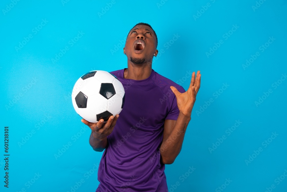 Man wearing purple T-shirt holding a ball over blue background crying and screaming. Human emotions, facial expression concept. Screaming, hate, rage. Man wearing purple T-shirt holding a ball over blue background crying and screaming. Human emotions, facial expression concept. Screaming, hate, rage.