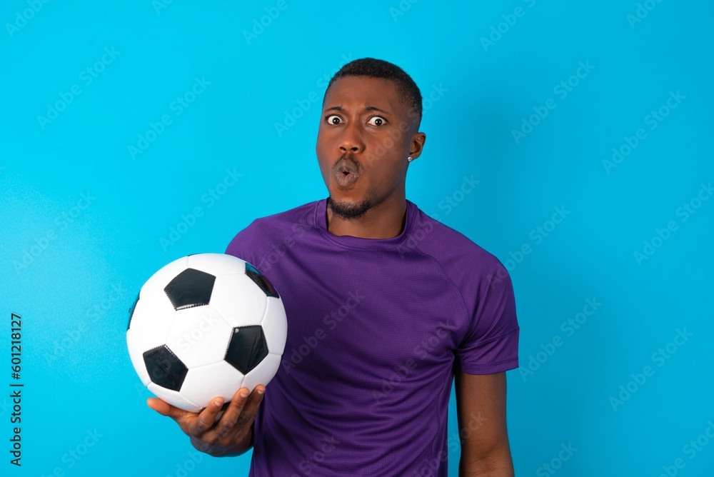 Man wearing purple T-shirt holding a ball over blue background expressing disgust, unwillingness, disregard having tensive look frowning face, looking indignant with something. Man wearing purple T-shirt holding a ball over blue background expressing disgust, unwillingness, disregard having tensive look frowning face, looking indignant with something.