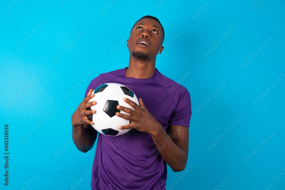 Happy smiling Man wearing purple T-shirt holding a ball over blue background has hands on chest near heart. Human emotions, real feelings and facial expression concept.