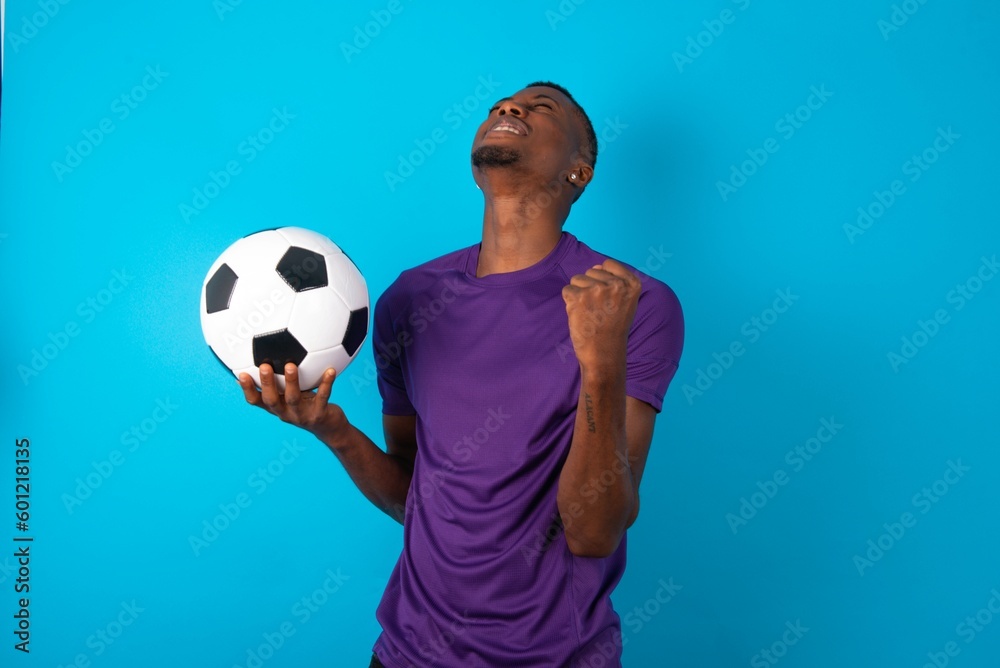 Man wearing purple T-shirt holding a ball over blue background looks with excitement up, keeps hands raised, notices something unexpected. Man wearing purple T-shirt holding a ball over blue background looks with excitement up, keeps hands raised, notices something unexpected.