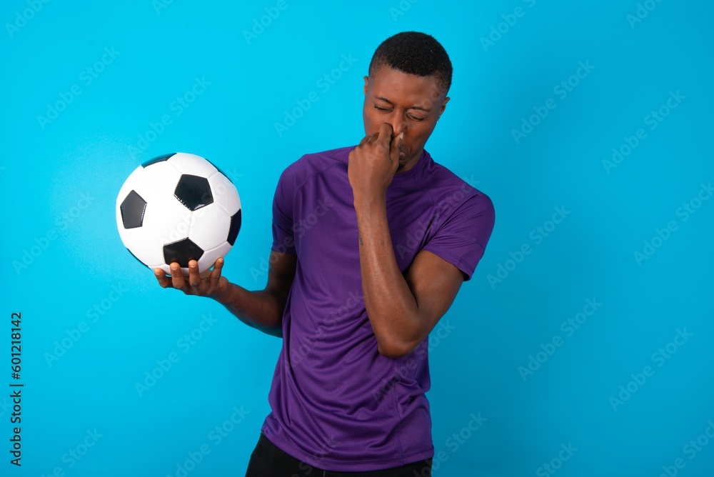 Man wearing purple T-shirt holding a ball over blue background , holding his nose because of a bad smell. Man wearing purple T-shirt holding a ball over blue background , holding his nose because of a bad smell.