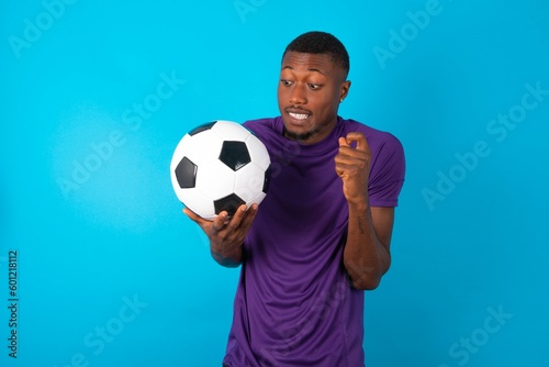 Portrait of desperate and shocked Man wearing purple T-shirt holding a ball over blue background looking panic, holding hands near face, with mouth wide open.