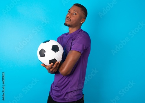 Image of cheerful Man wearing purple T-shirt holding a ball over blue background with arms crossed. Looking and smiling at the camera. Confidence concept.