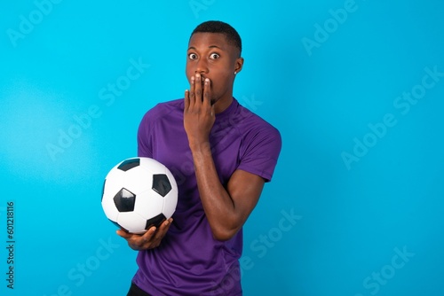 Oh! I think I said it! Close up portrait Man wearing purple T-shirt holding a ball over blue background cover open mouth by hand palm, look at camera with big eyes.