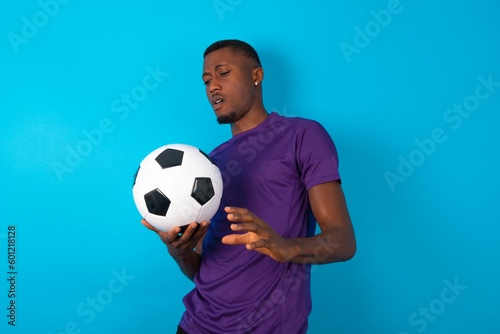 Ugh how disgusting! Displeased Man wearing purple T-shirt holding a ball over blue background , has dissatisfied facial expression as sees something abominable.