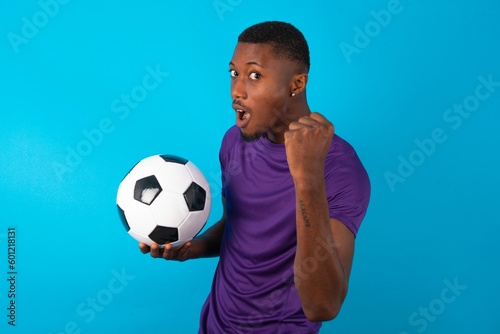 Fierce Man wearing purple T-shirt holding a ball over blue background holding fist in front as if is ready for fight or challenge, screaming and having aggressive expression on face.