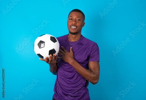 Honest Man wearing purple T-shirt holding a ball over blue background keeps hands on chest, touched by compliment or makes promise, looks at camera with great pleasure.