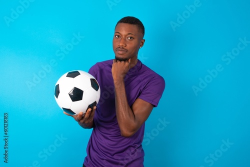 Man wearing purple T-shirt holding a ball over blue background with hand under chin and looking sideways with doubtful and skeptical expression, suspect and doubt.