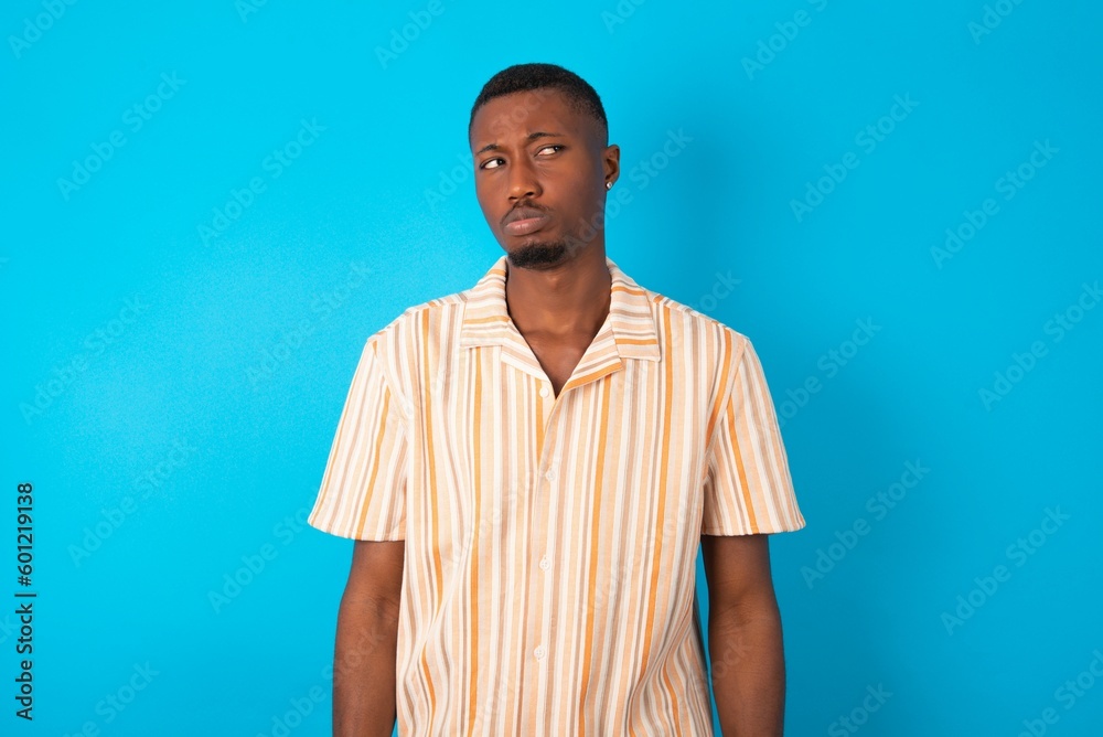 Dissatisfied Handsome man wearing fashion shirt over blue background purses lips and has unhappy expression looks away stands offended. Depressed frustrated model. Dissatisfied Handsome man wearing fashion shirt over blue background purses lips and has unhappy expression looks away stands offended. Depressed frustrated model.