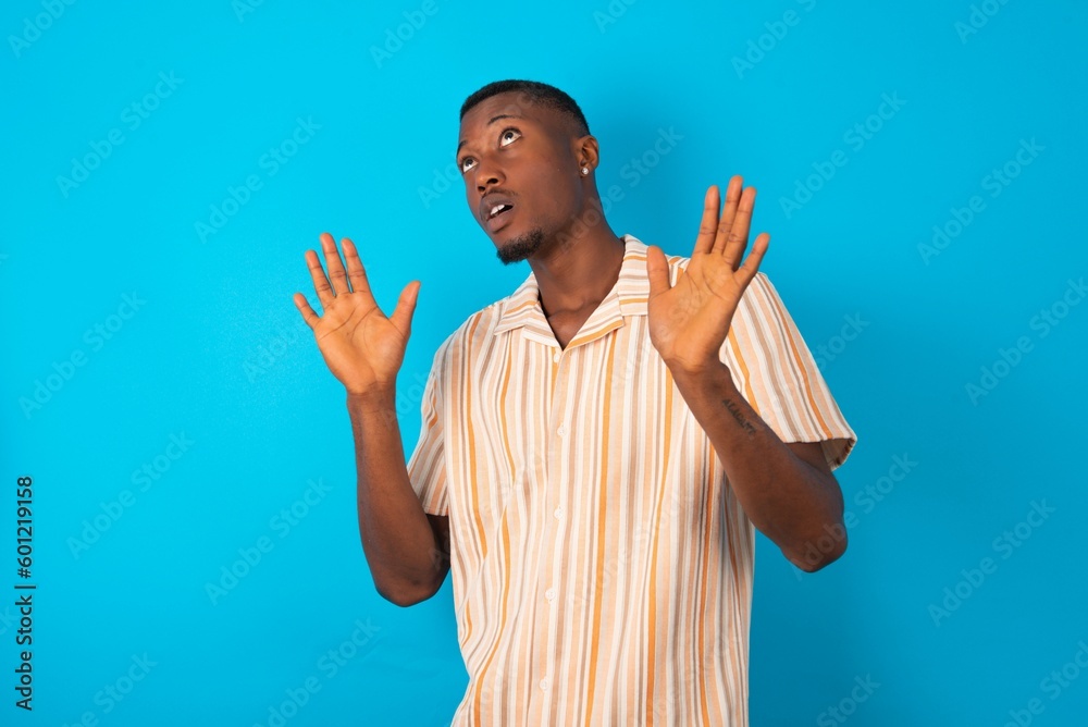 Handsome man wearing fashion shirt over blue background keeps palms forward and looks with fright above on ceiling tries to defense herself from invisible danger opens mouth. Handsome man wearing fashion shirt over blue background keeps palms forward and looks with fright above on ceiling tries to defense herself from invisible danger opens mouth.