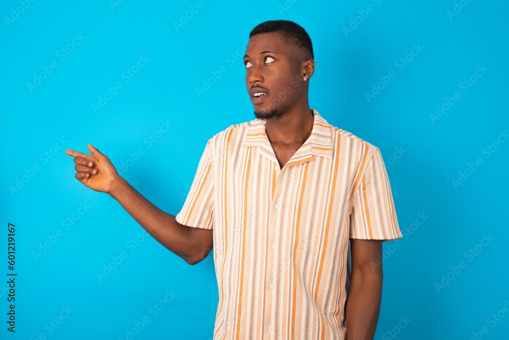 Handsome man wearing fashion shirt over blue background points at copy space and advertises something, advices best price. Handsome man wearing fashion shirt over blue background points at copy space and advertises something, advices best price.