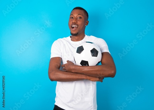 Young man wearing white T-shirt holding a ball over blue background being happy smiling and crossed arms looking confident at the camera. Positive and confident person.