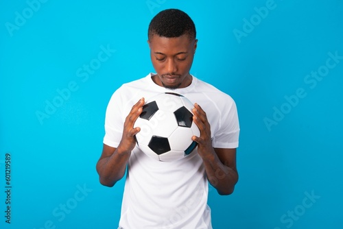 Indoor closeup of Young man wearing white T-shirt holding a ball over blue background practicing yoga and meditation, holding palms together in namaste, looking calm, relaxed and peaceful.