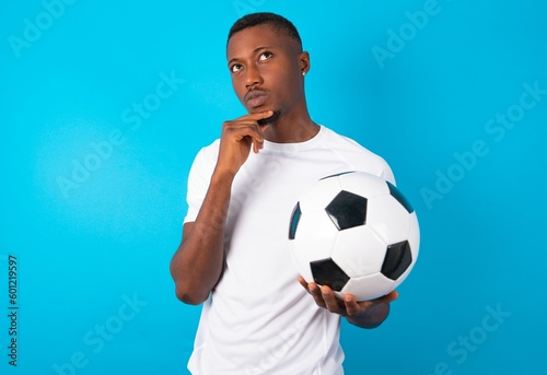 Shot of contemplative thoughtful Young man wearing white T-shirt holding a ball over blue background keeps hand under chin, looks thoughtfully upwards.
