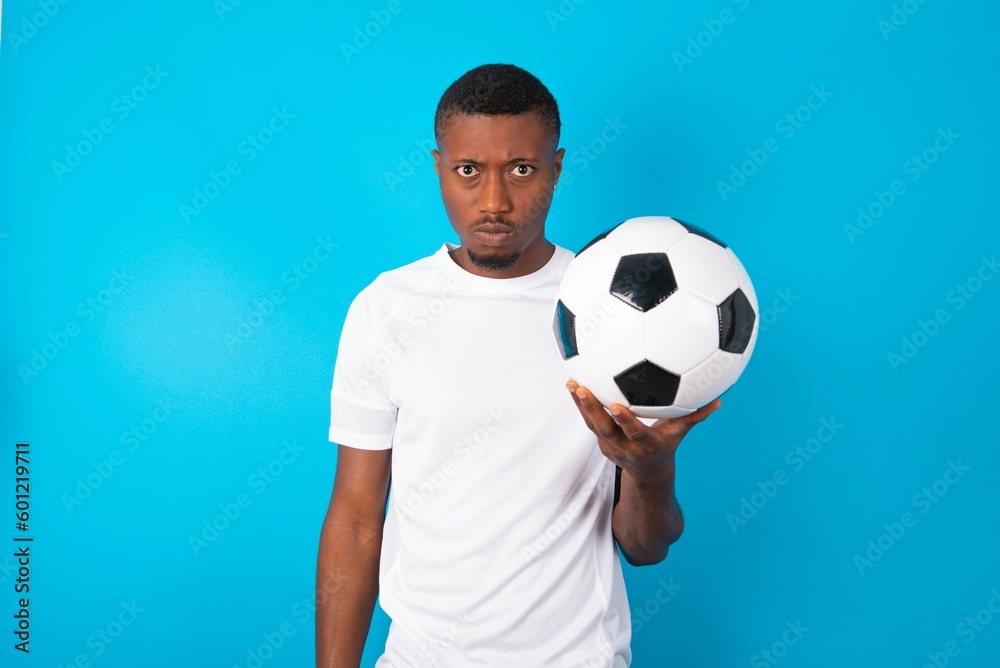Young man wearing white T-shirt holding a ball over blue background frowning his eyebrows being displeased with something. Young man wearing white T-shirt holding a ball over blue background frowning his eyebrows being displeased with something.