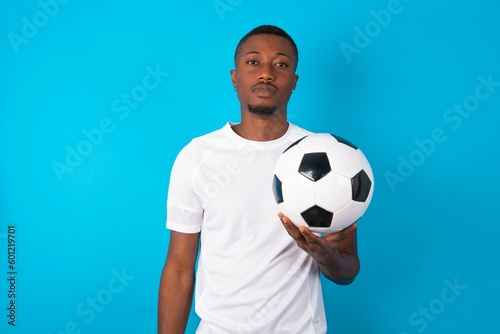 Joyful Young man wearing white T-shirt holding a ball over blue background looking to the camera, thinking about something. Both arms down, neutral facial expression.