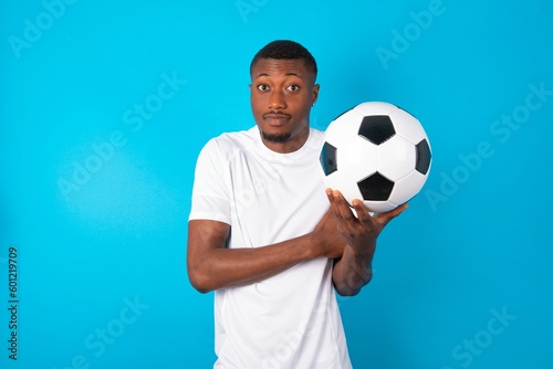 Young man wearing white T-shirt holding a ball over blue background bitting his mouth and looking worried and scared crossing arms, worry and doubt.