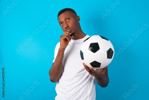 Portrait of thoughtful Young man wearing white T-shirt holding a ball over blue keeps hand under chin, looks away trying to remember something or listens something with interest. Youth concept.
