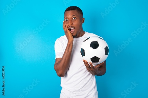 Shocked, astonished Young man wearing white T-shirt holding a ball looking surprised in full disbelief wide open mouth with hand near face. Positive emotion facial expression body language.