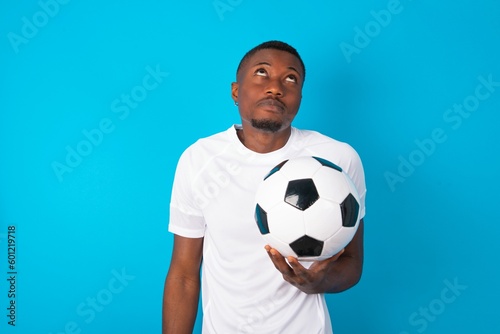 Portrait of mysterious Young man wearing white T-shirt holding a ball over blue background looking up with enigmatic smile. Advertisement concept.