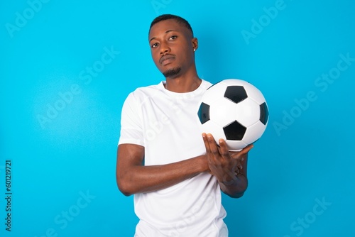 Cheerful Young man wearing white T-shirt holding a ball over blue background with hand near face. Looking with glad expression at the camera after listening to good news. Confidence.