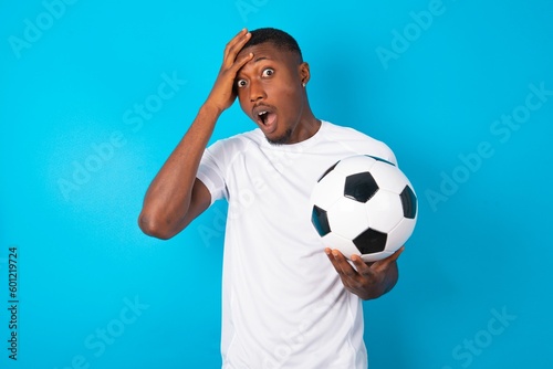 Shocked panic Young man wearing white T-shirt holding a ball over blue background holding hands on head and screaming in despair and frustration.