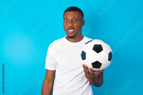 Mad crazy Young man wearing white T-shirt holding a ball over blue background clenches teeth angrily, being annoyed with coming noise. Negative feeling concept.