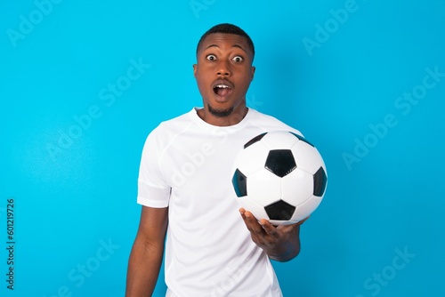 Expressive facial expressions. Shocked stupefied Young man wearing white T-shirt holding a ball over blue background , keeps jaw dropped feels stunned from what he sees aside.