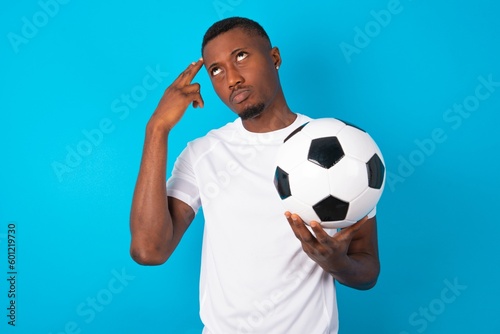 Unhappy Young man wearing white T-shirt holding a ball over blue background imitates gun shoot makes suicide gesture keeps two fingers on temples.