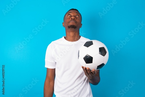 Young man wearing white T-shirt holding a ball over blue background looking up as he sees something strange.