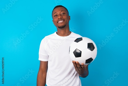 Young man wearing white T-shirt holding a ball over blue background with broad smile, shows white teeth, feeling confident rejoices having day off.