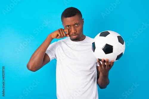 Unhappy Young man wearing white T-shirt holding a ball over blue background crying while posing at camera whipping tears with hand.
