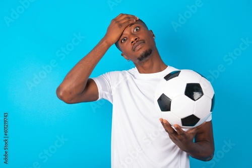 Oops, what did I do? Young man wearing white T-shirt holding a ball over blue background holding hand on forehead with frightened and regret expression.