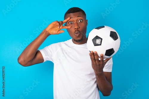 Young man wearing white T-shirt holding a ball over blue background making v-sign near eyes. Leisure lifestyle people person celebrate flirt coquettish concept.