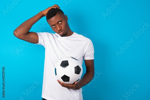 Young man wearing white T-shirt holding a ball over blue background saying: Oops, what did I do? Holding hand on head with frightened and regret expression.