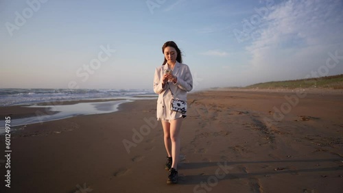 A young woman in a white jacket and black shorts, walking along the windy shoreline at sunset on a beach in Uchinada Town, Ishikawa Prefecture. photo