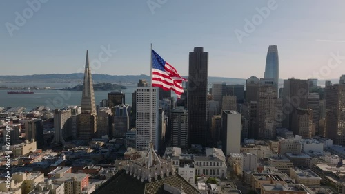 Slide and pan footage of US flag waving in wind on top of building above city. Modern downtown skyscrapers in background. San Francisco, California, USA