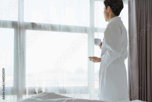 Tourist Asian man hold coffee cup in bathrobe towel looking at beautiful scene view on window at hotel room. Traveler Asian man feels relax in travel on holiday trip. Tourist relax in hotel concept.