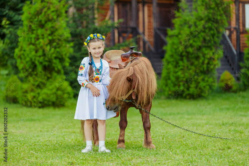 Obraz premium young girl in Ukrainian national dress strokes a pony that grazes on the lawn
