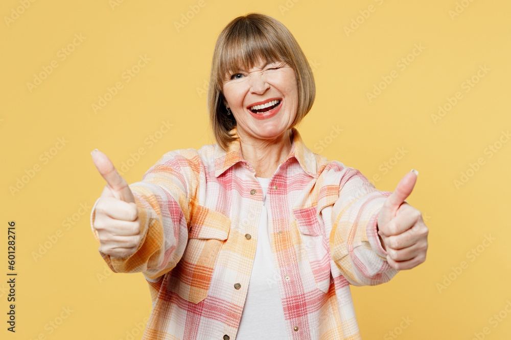 Elderly smiling happy blonde caucasian woman 50s years old she wear casual clothes showing thumb up like gesture wink blink eye isolated on plain yellow background studio portrait. Lifestyle concept.