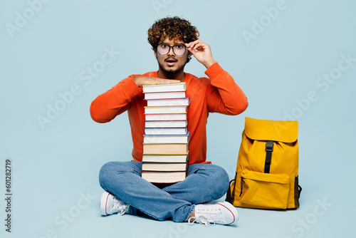 Full body shocked young teen Indian boy student wear casual clothes glasses sit near backpack bag hold pile stack of many books isolated on plain pastel blue background. High school college concept. © ViDi Studio