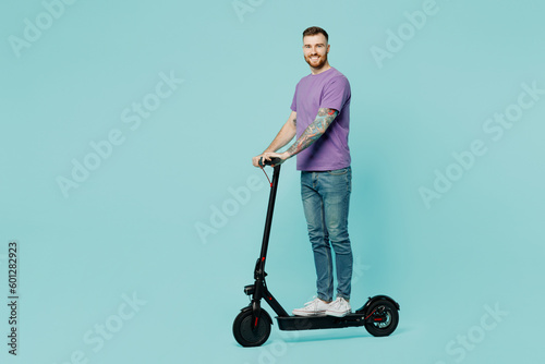 Full body sideways caucasian smiling fun happy young man he wearing purple t-shirt riding e-scooter look camera isolated on plain pastel light blue cyan background studio portrait. Lifestyle concept.