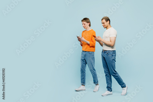 Full body young couple two gay men wear casual clothes together hold use mobile cell phone go isolated on pastel plain blue color background studio portrait. Pride day june month love LGBTQ concept.