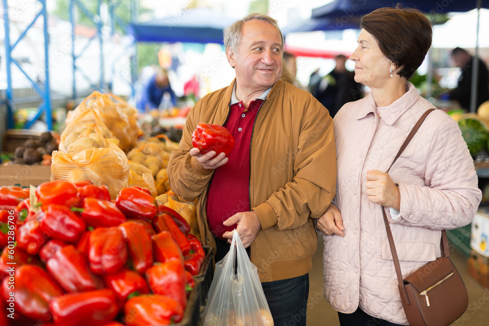 Positive senior couple in casual wear picking bell peppers during shopping at crowded bazaar