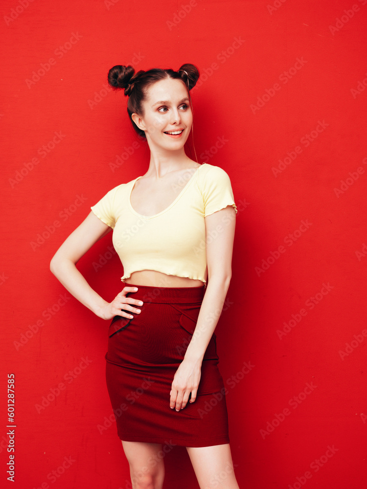 Young beautiful smiling female in trendy summer yellow t-shirt and skirt. Carefree woman with two horns hairstyle posing near red wall in studio. Positive model having fun. Cheerful and happy Young beautiful smiling female in trendy summer yellow t-shirt and skirt. Carefree woman with two horns hairstyle posing near red wall in studio. Positive model having fun. Cheerful and happy