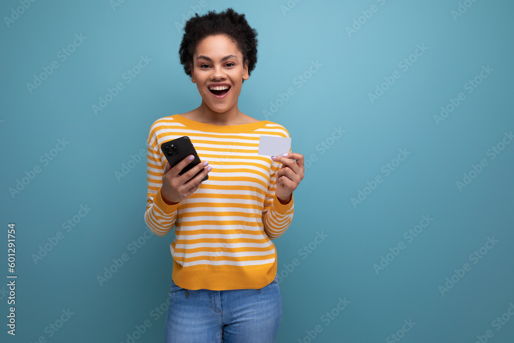 joyful latin young woman with black afro hair with phone and money card on background with copy space joyful latin young woman with black afro hair with phone and money card on background with copy space
